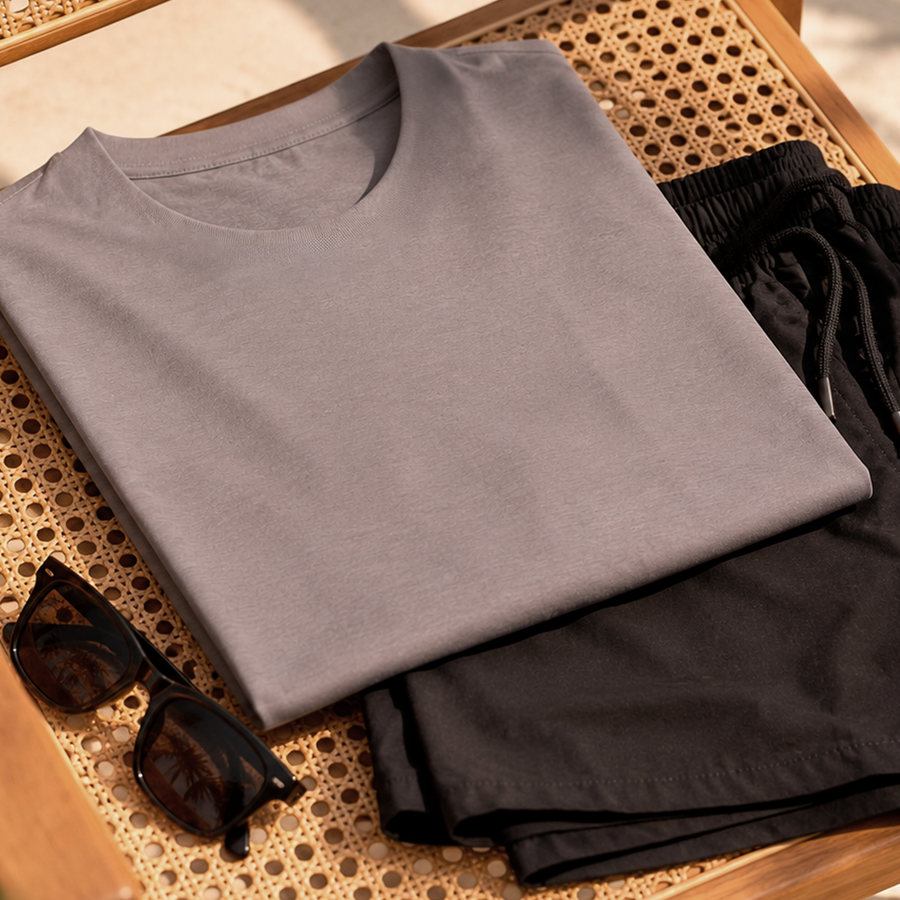 Camiseta cinza dobrada, bermuda preta e óculos de sol sobre superfície de palhinha, look masculino leve para dias de calor